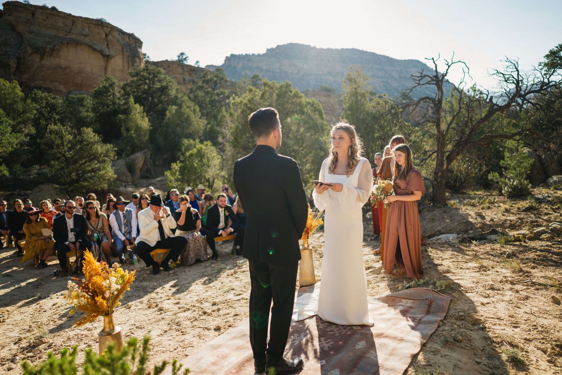 20210905_BrianTessWedding_429 A couple exchanges vows during an outdoor wedding ceremony held in a sunlit, rocky landscape with guests seated nearby.
