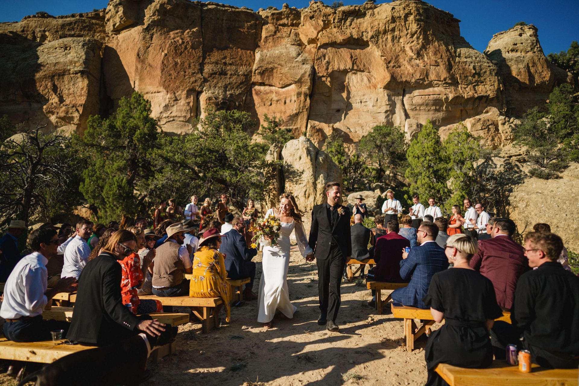 20210905_BrianTessWedding_485 A newlywed couple walks hand-in-hand down an aisle of wooden benches at an outdoor desert wedding near rocky cliffs.