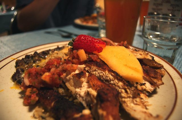 A plate of sliced grilled chicken topped with a mango slice and a strawberry, served in a restaurant setting.