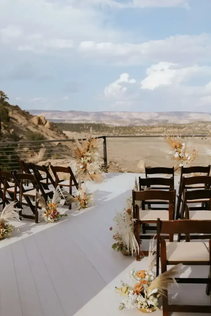 Outdoor wedding ceremony setup with rows of wooden chairs, floral arrangements, and a scenic desert landscape background.