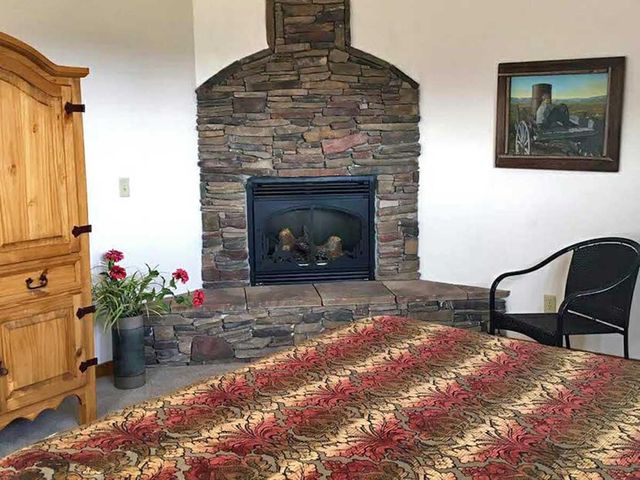 A bedroom corner with a stone fireplace, a tall wooden armoire, a patterned quilt on a bed, and a black metal chair.