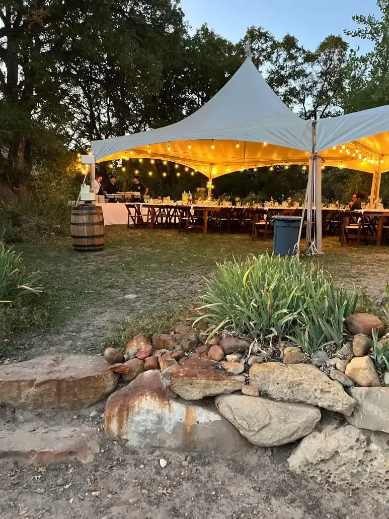 A white event tent illuminated by string lights at dusk, set over dining tables on a grass lawn near rocky landscaping.