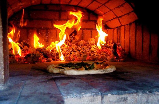 A pizza cooks on a stone surface inside a brick oven with bright orange flames burning in the background.