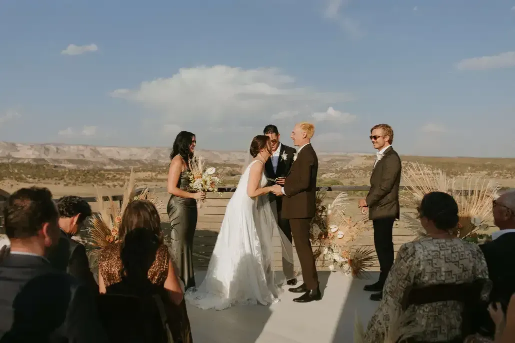 A wedding ceremony takes place outdoors in a desert landscape, featuring a couple standing before an officiant.