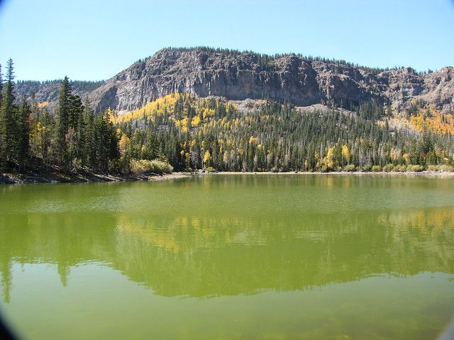 A calm green lake reflecting a forested mountainside with yellow autumn foliage under a clear blue sky.