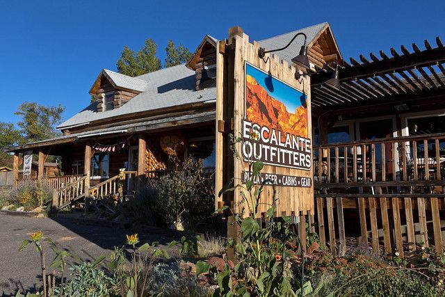 A rustic wooden Escalante Outfitters shop building under a clear blue sky, with a prominent branded sign in the foreground.