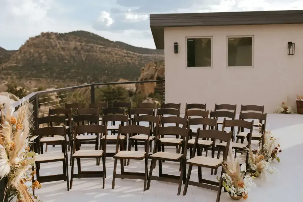 Rows of brown wooden folding chairs with beige cushions arranged on a deck for an outdoor wedding with mountain views.