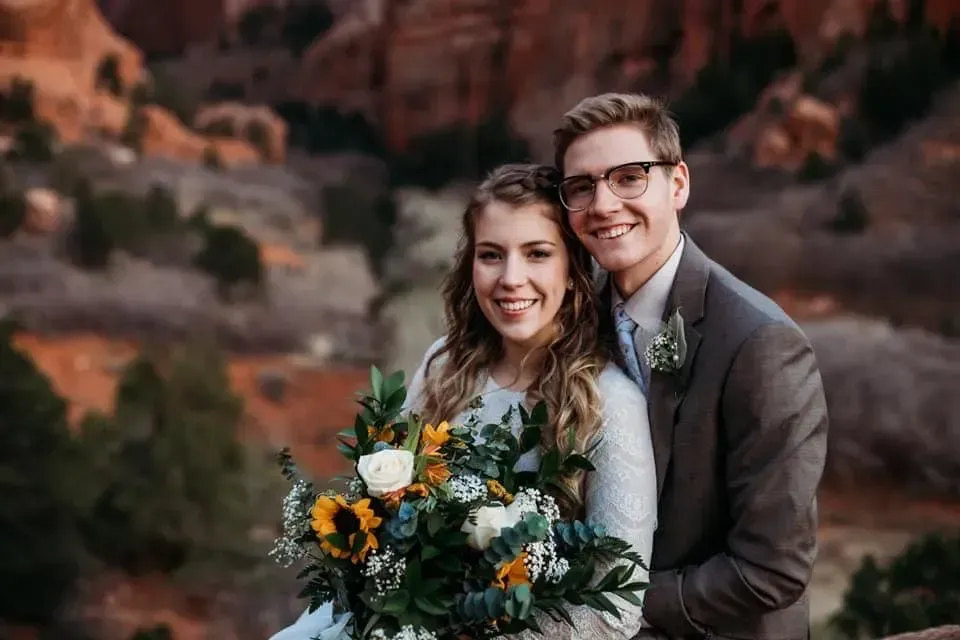 A smiling couple in formal wedding attire holds a wildflower bouquet against a backdrop of red desert rock formations.