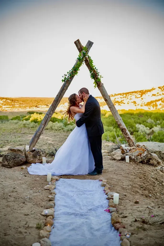 A couple kisses under a rustic, triangular wooden arch decorated with greenery, atop a hill with a dirt aisle below.