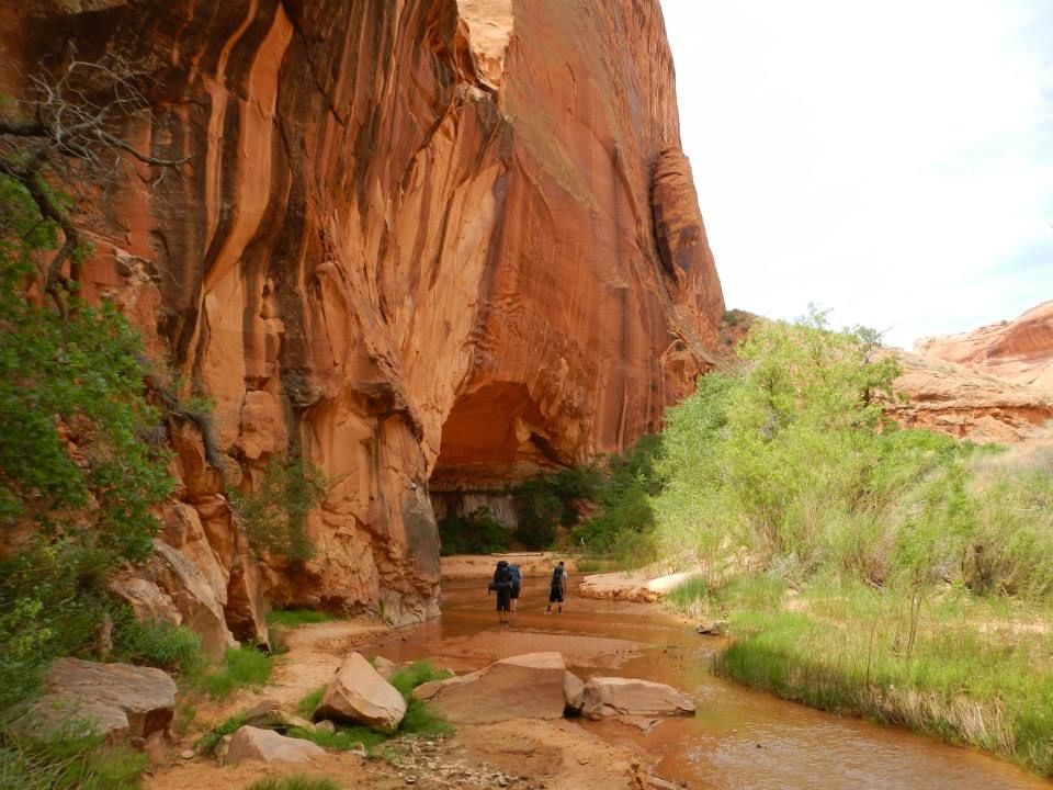 Two people with backpacks hike along a shallow, sandy stream at the base of a towering, reddish-orange sandstone cliff.