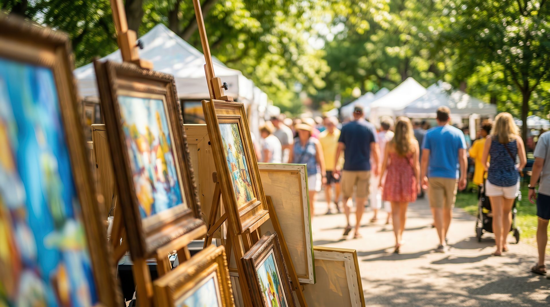 Framed paintings displayed outdoors at an art fair with people walking down a sunlit path