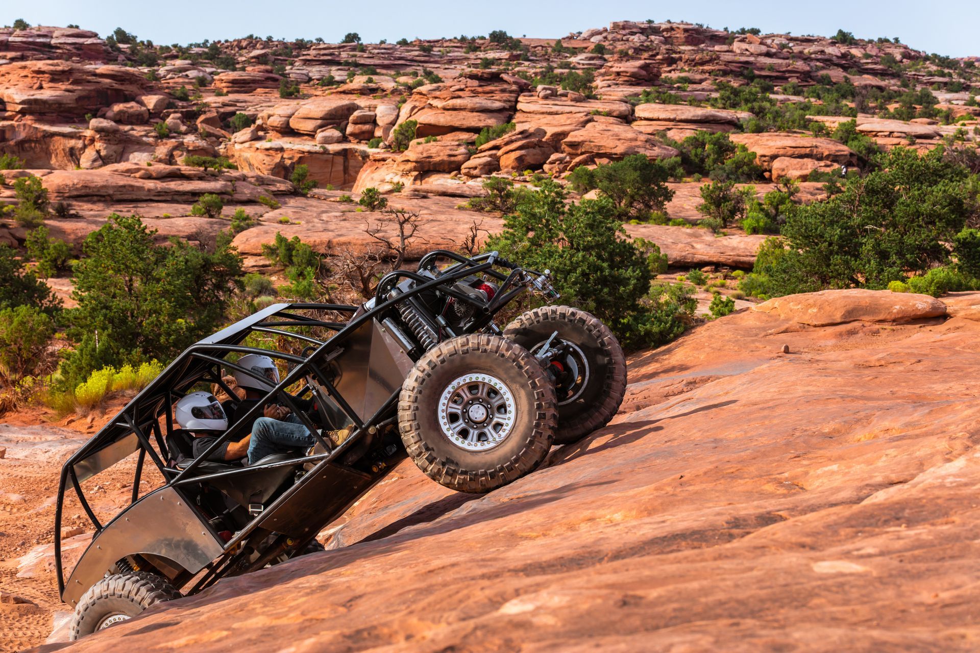 A black off-road vehicle climbs a steep red rock hill in a rugged desert landscape under a clear sky.