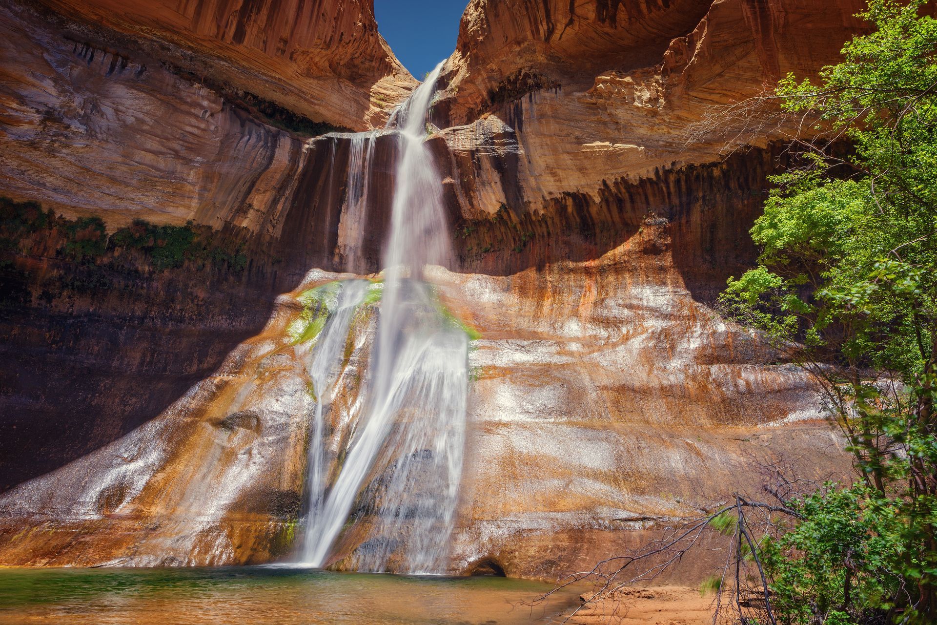 Waterfall cascading over orange-red canyon cliffs into a shallow pool with green foliage nearby