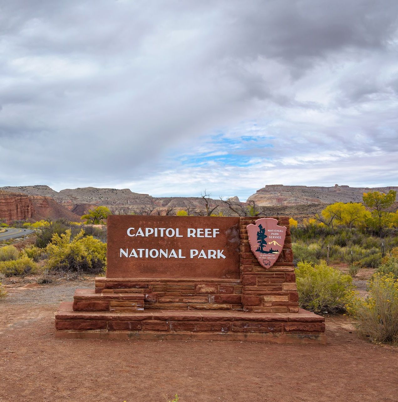 A red stone sign for Capitol Reef National Park against a backdrop of desert shrubs and mesas under a cloudy sky.