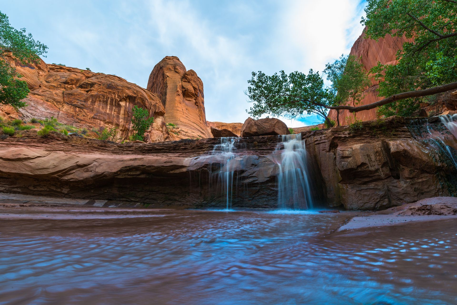A small waterfall cascades over red rock ledges into a stream, surrounded by desert cliffs and sparse green trees.