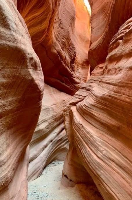 A narrow, winding slot canyon with smooth, wavy orange sandstone walls and a sandy floor under soft overhead light.