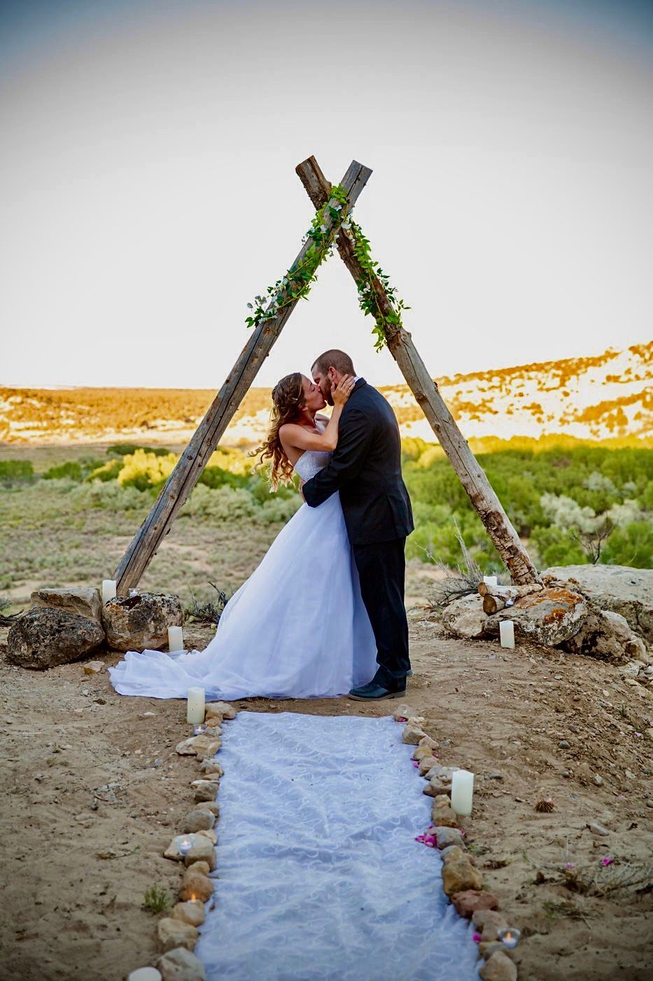 IMG_1968 A couple in wedding attire kisses under a rustic, triangular wooden arbor decorated with greenery on a desert hillside.