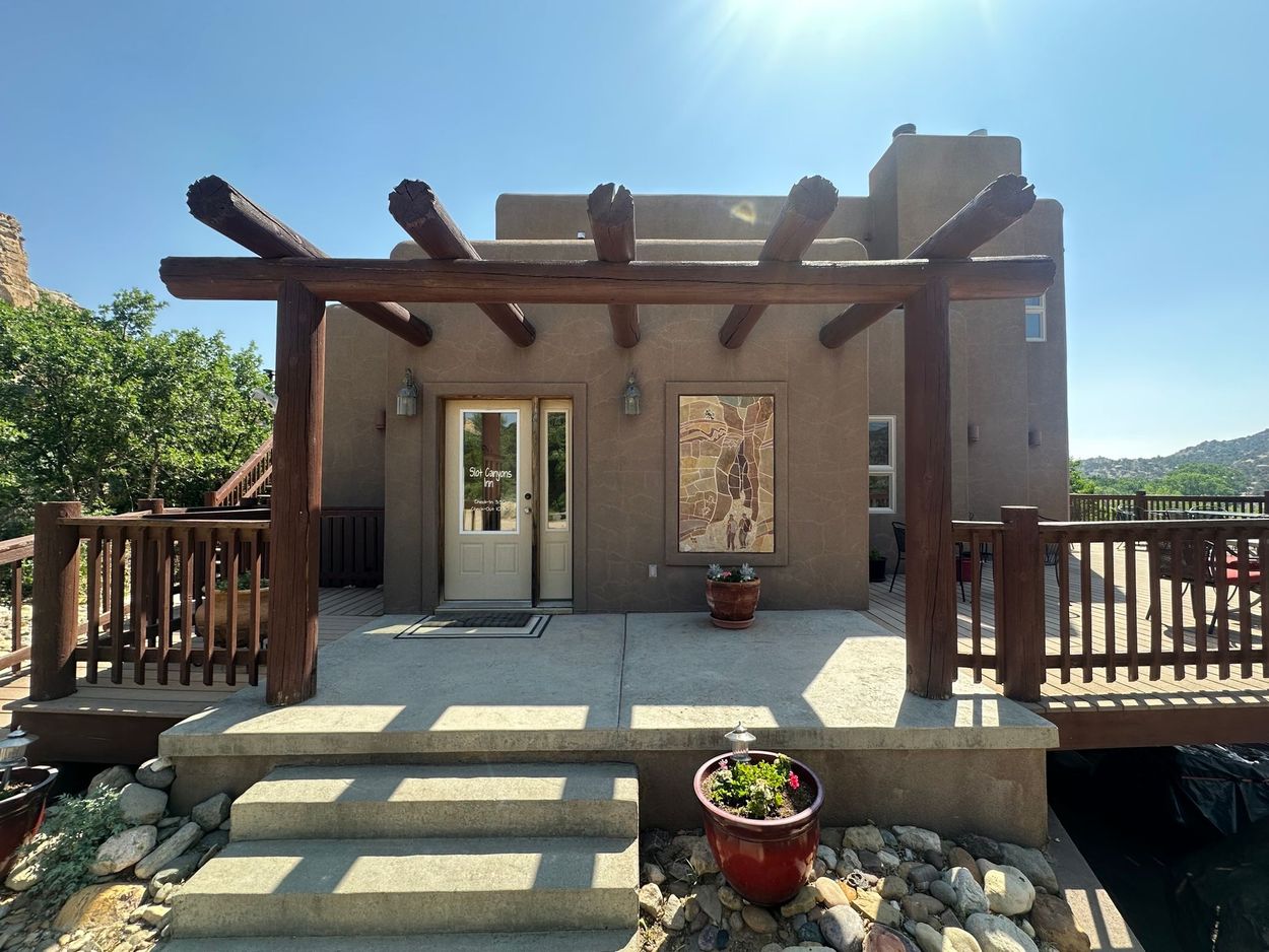 A tan stucco house with a wooden pergola over a concrete patio, stone steps, and a brown wooden deck railing.