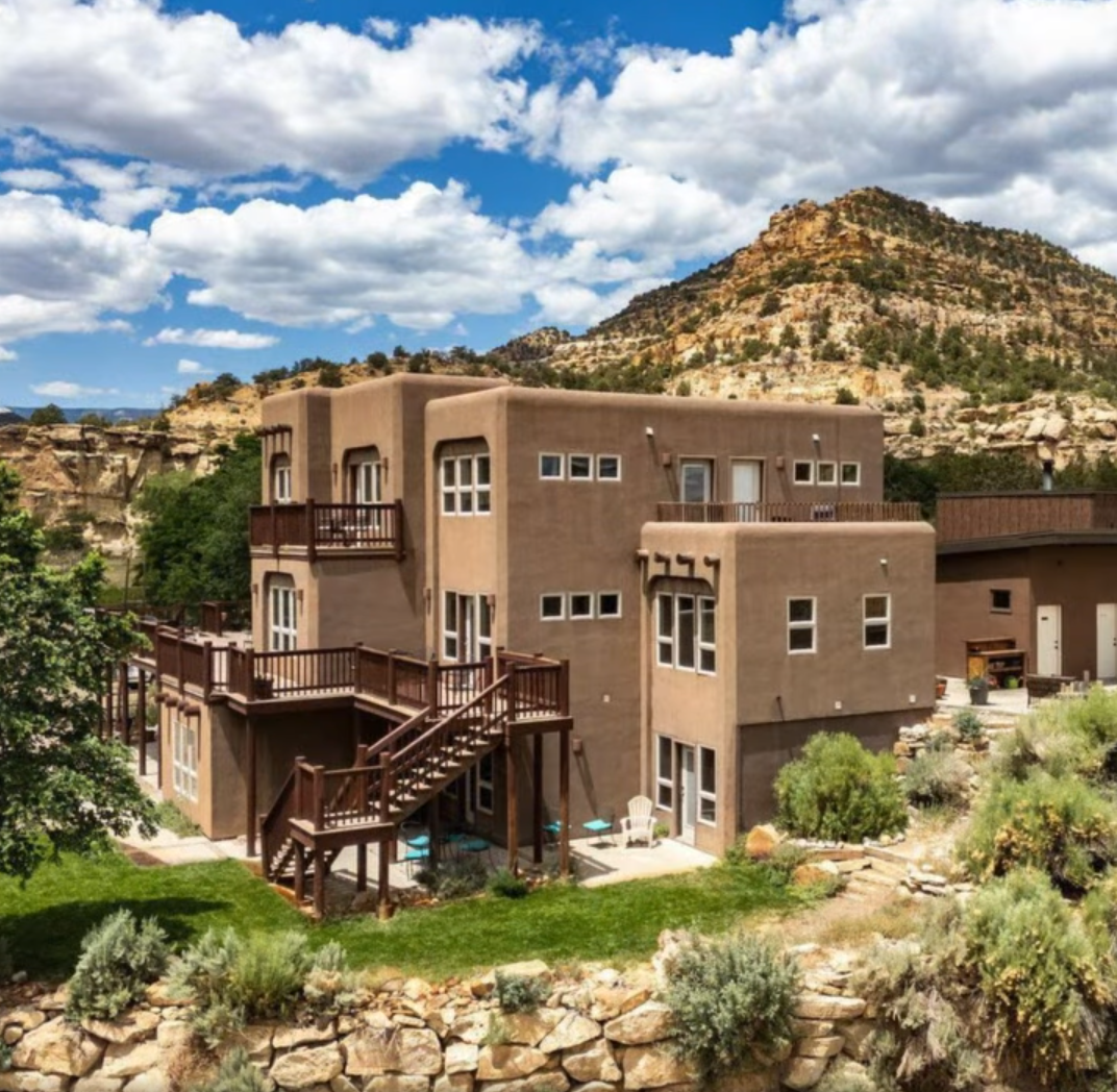 A multi-story, tan adobe-style house with wooden decks sits against a rugged, rocky mountain landscape under a blue sky.