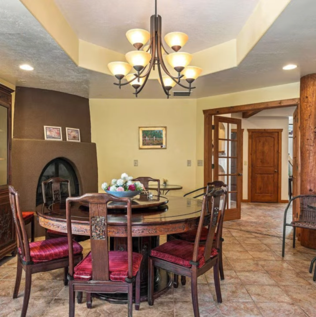A circular wooden dining table with chairs and a centerpiece sits under a chandelier in a room with a kiva-style fireplace.