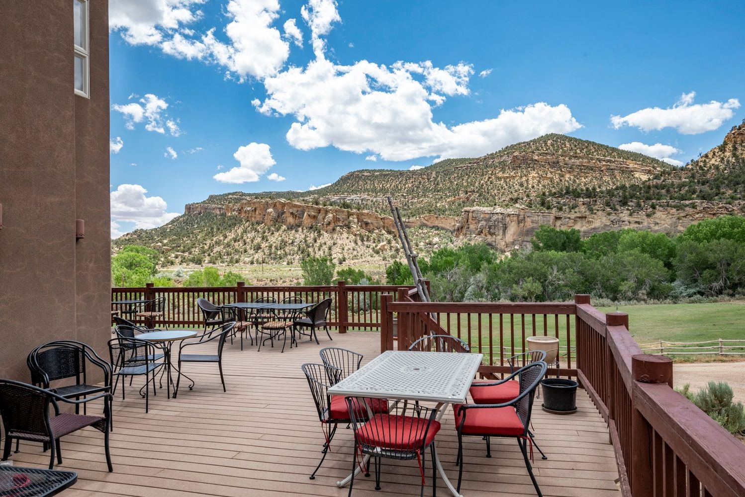 A spacious outdoor deck with tables and chairs, overlooking a green landscape and rocky mountains under a sunny sky.