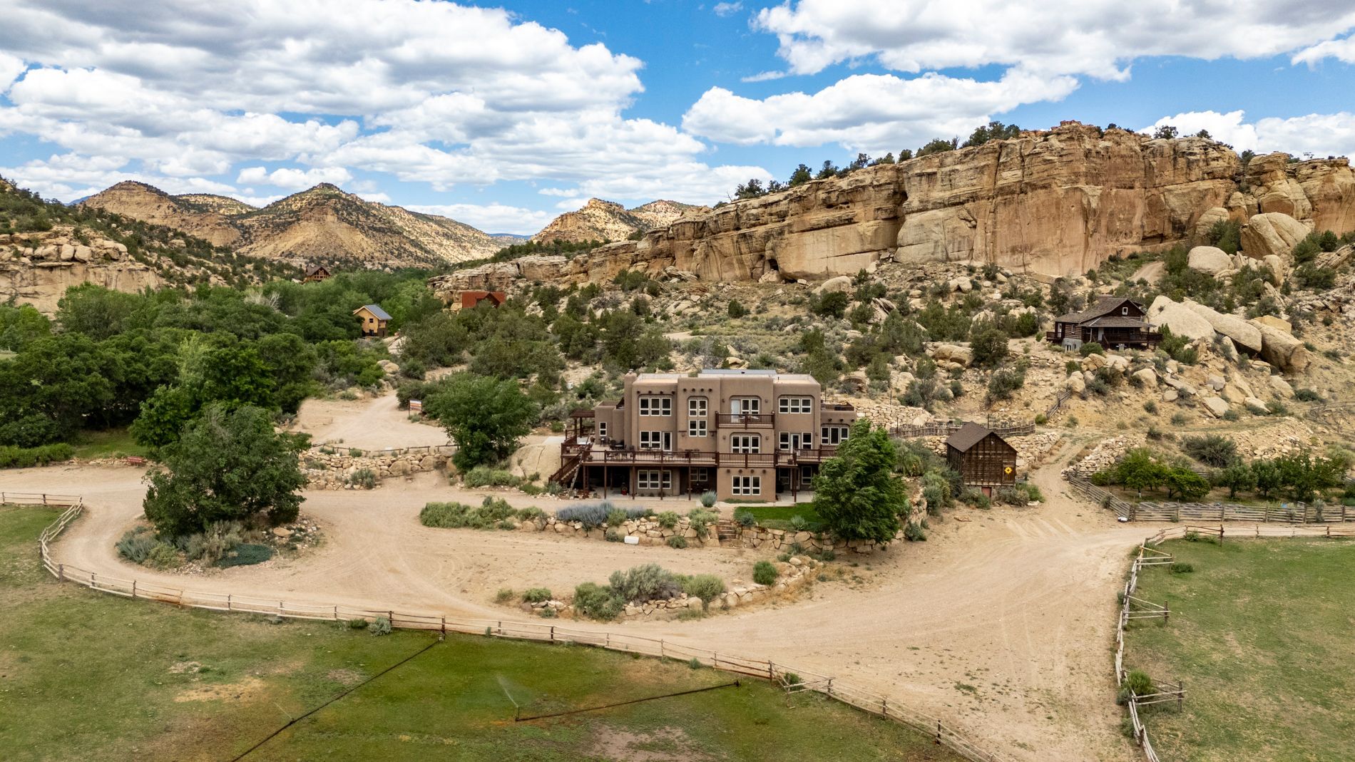 An aerial view of a multi-story brown house nestled against a rocky cliffside in a dry, desert landscape.