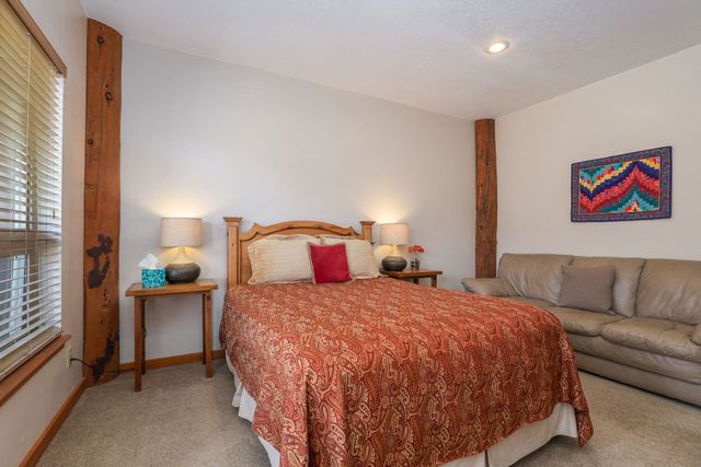 A bedroom featuring a bed with a patterned orange quilt, a side table with a lamp, and a tan sofa against a wall.