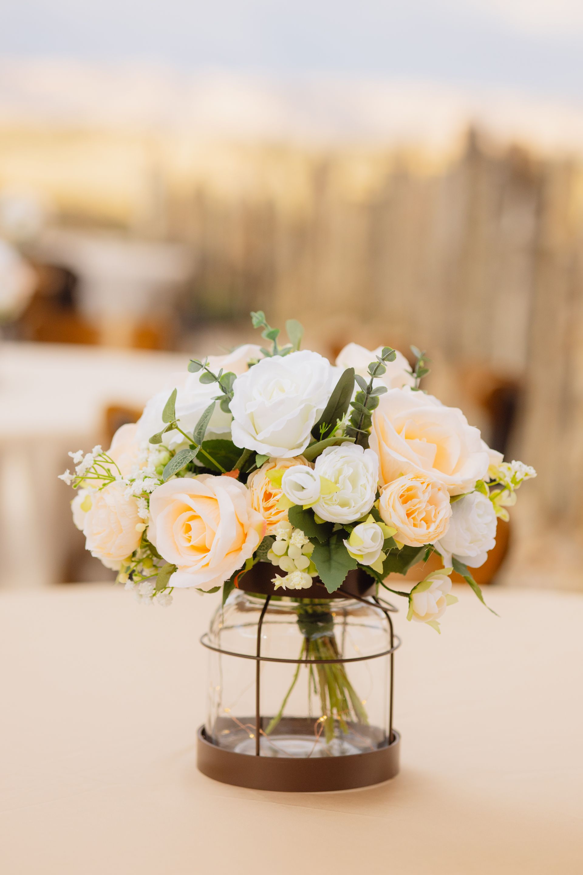 A bouquet of white and peach roses in a small, wire-caged glass lantern vase sitting on a neutral table.