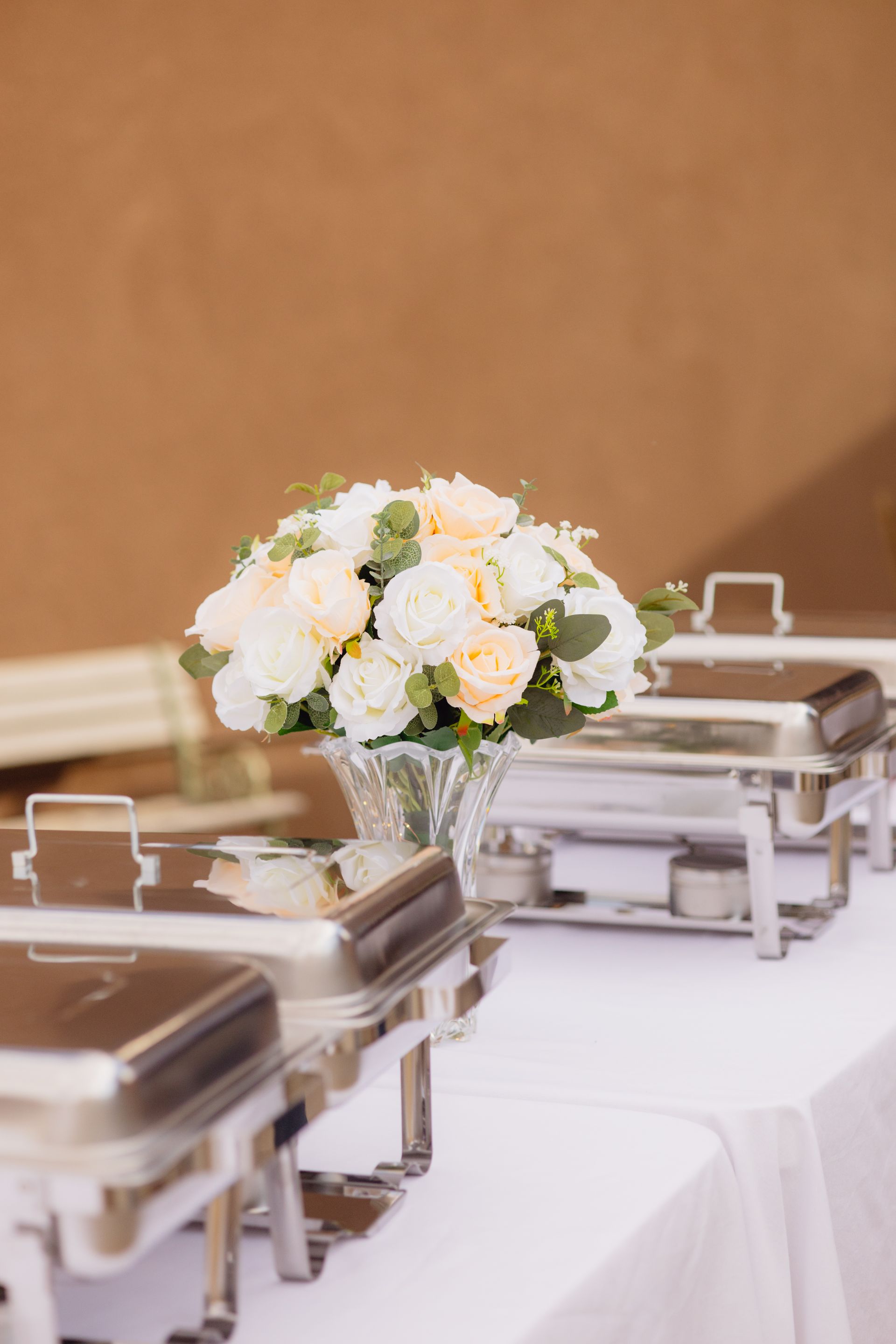 A crystal vase of white and pale peach roses sits on a white-covered banquet table between two stainless steel chafers.