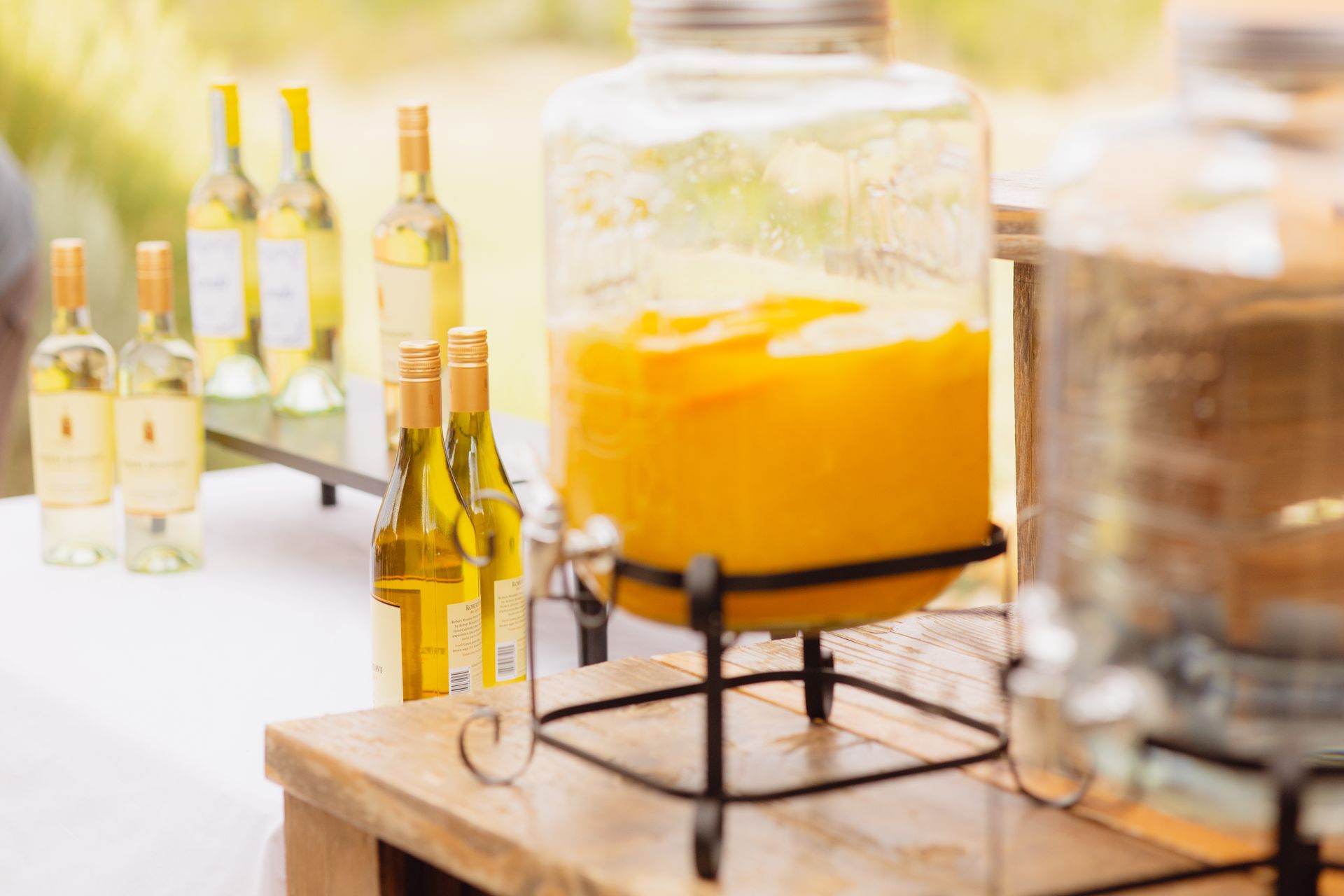 Glass dispensers of yellow and clear beverages on a wooden table, flanked by multiple wine bottles.