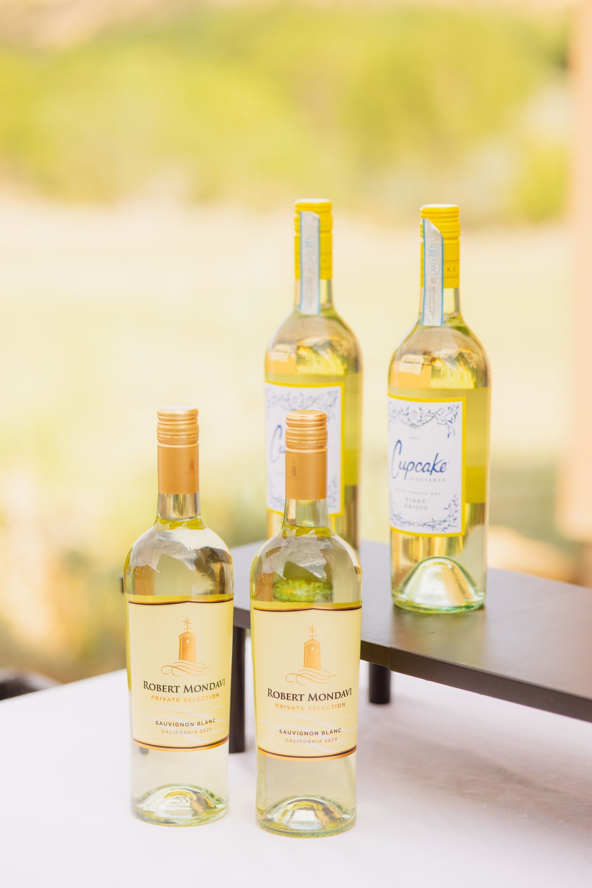 Four white wine bottles, two in the foreground and two on a small riser, set on a white table against a blurry background.
