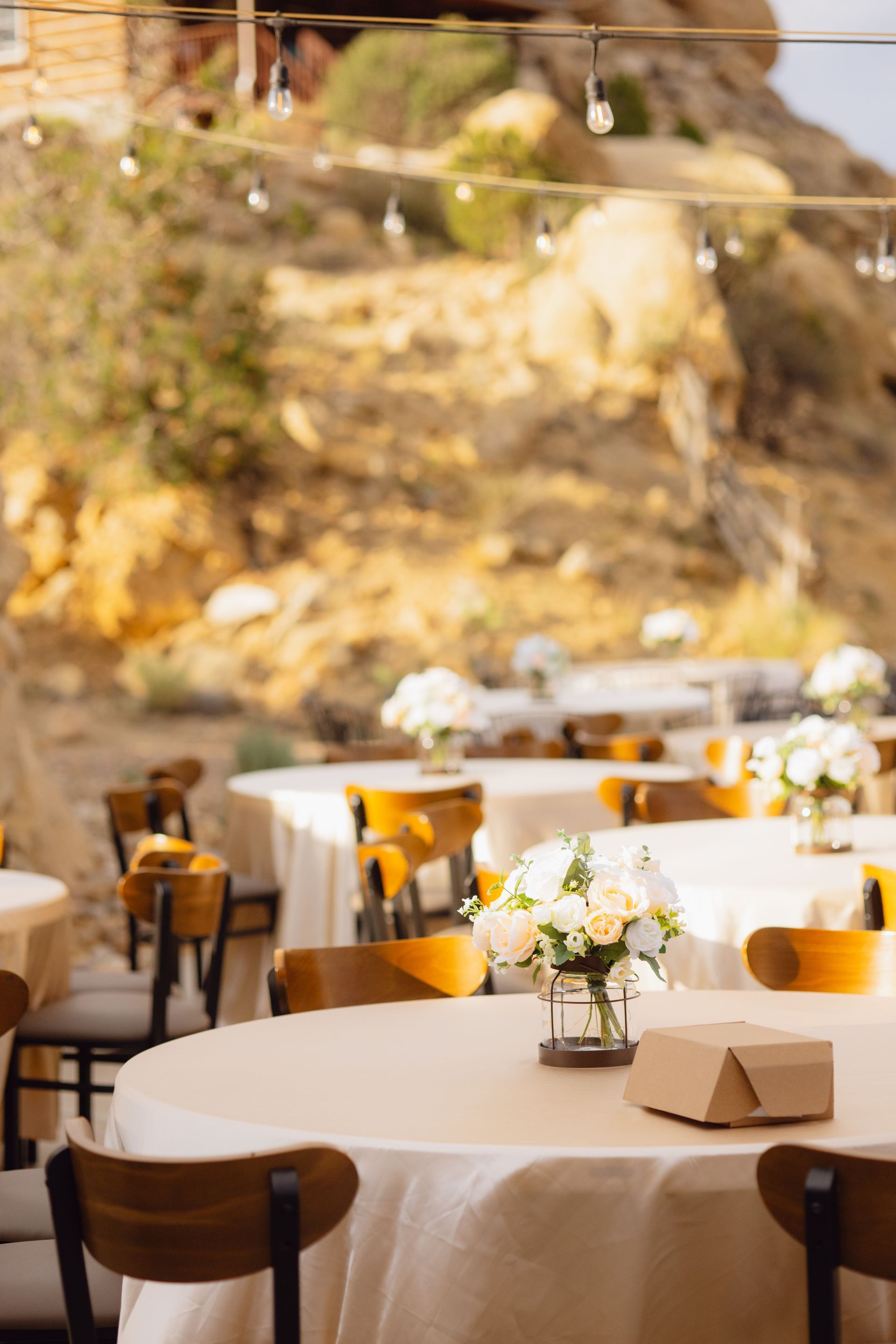 Outdoor wedding reception with circular tables topped with white tablecloths and floral centerpieces under hanging lights.