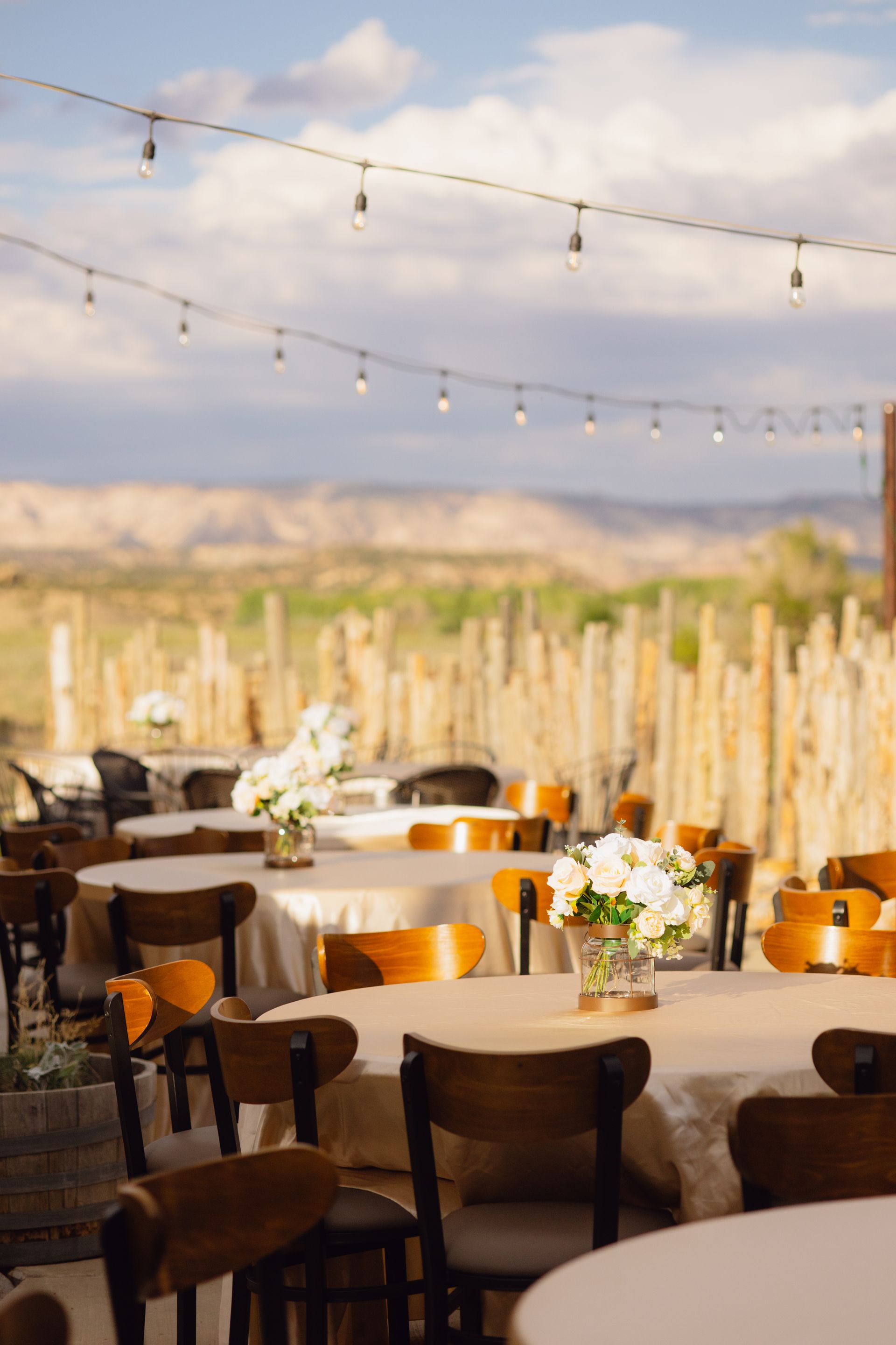 Outdoor dining setup with round tables and chairs under hanging string lights against a scenic desert landscape.