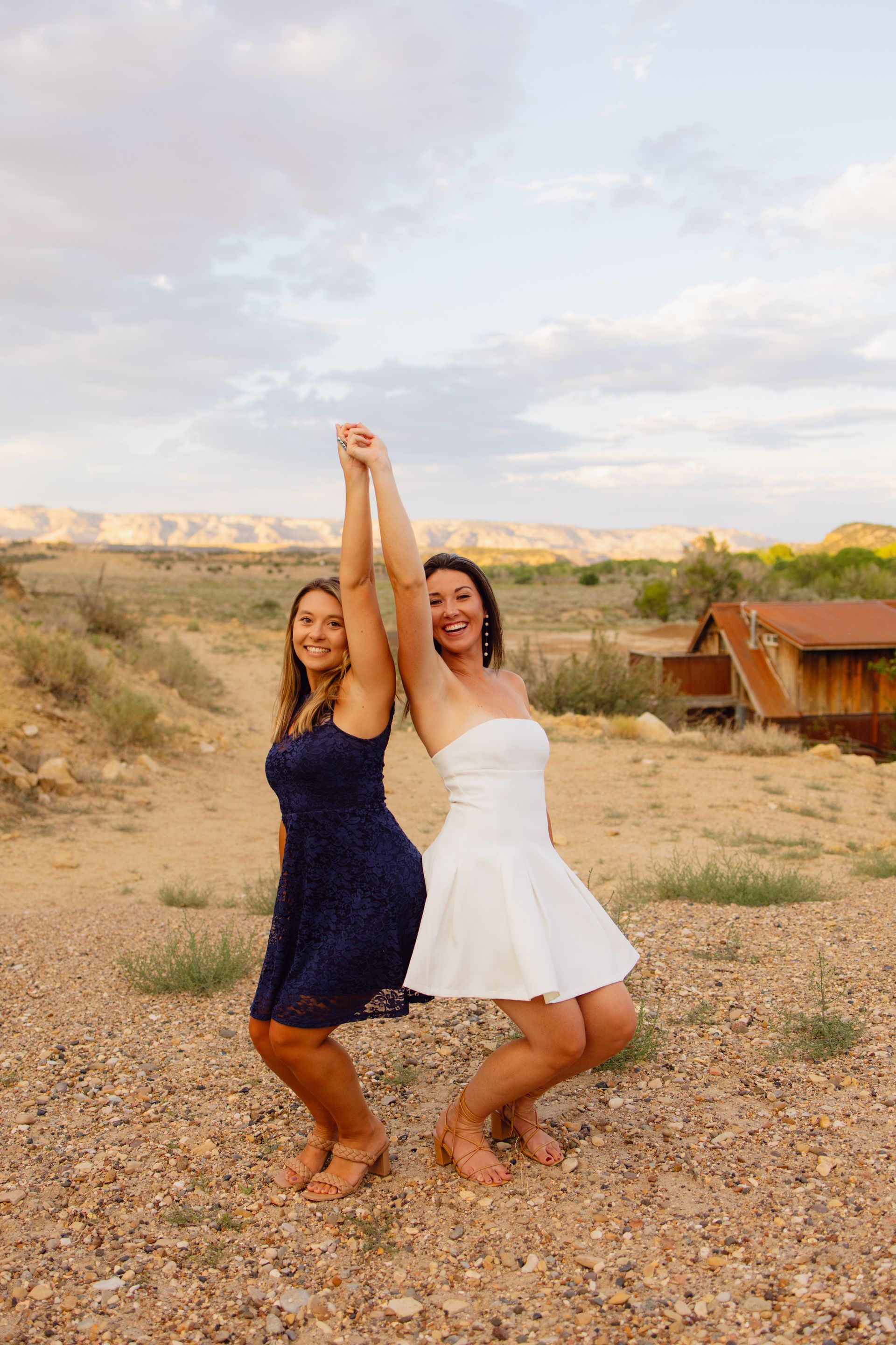 Two people in a dark blue and white dress pose back-to-back, holding hands raised in an outdoor desert setting.