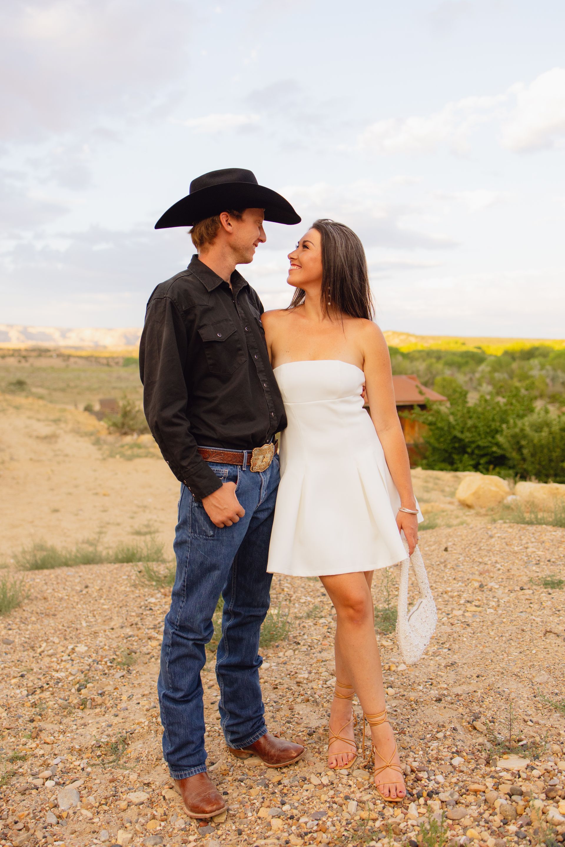 A couple in western attire standing in a field looking at each other under a partly cloudy sky.