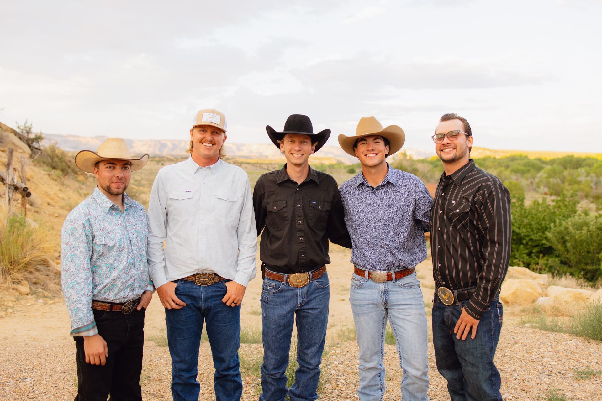 Five people in Western-style attire, including cowboy hats, shirts, and jeans, stand together outdoors on a dirt path.