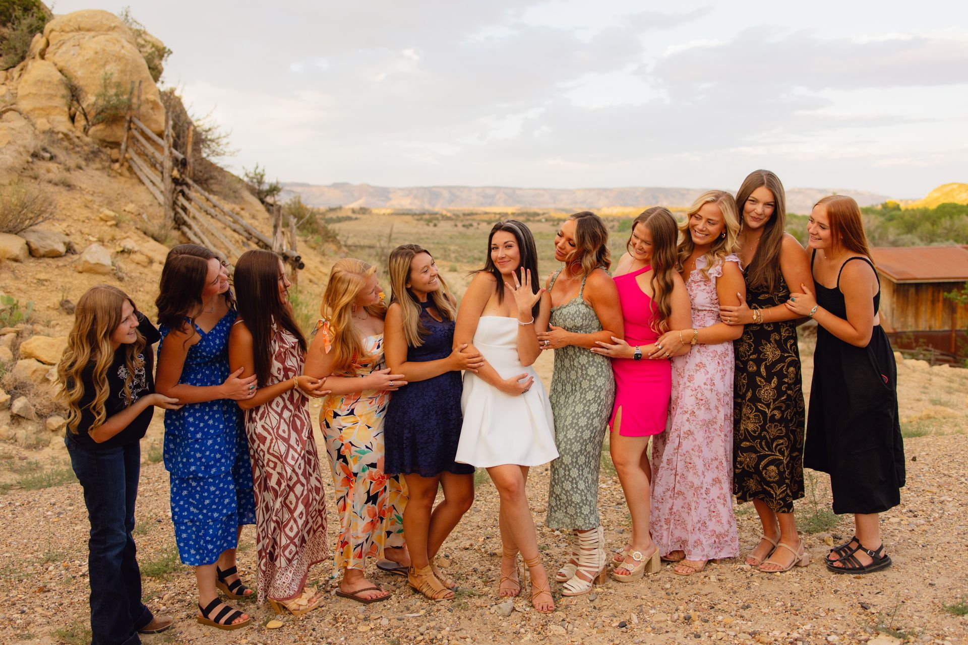 A group of friends stands in a line outdoors near a rocky hillside, laughing and showing off an engagement ring.
