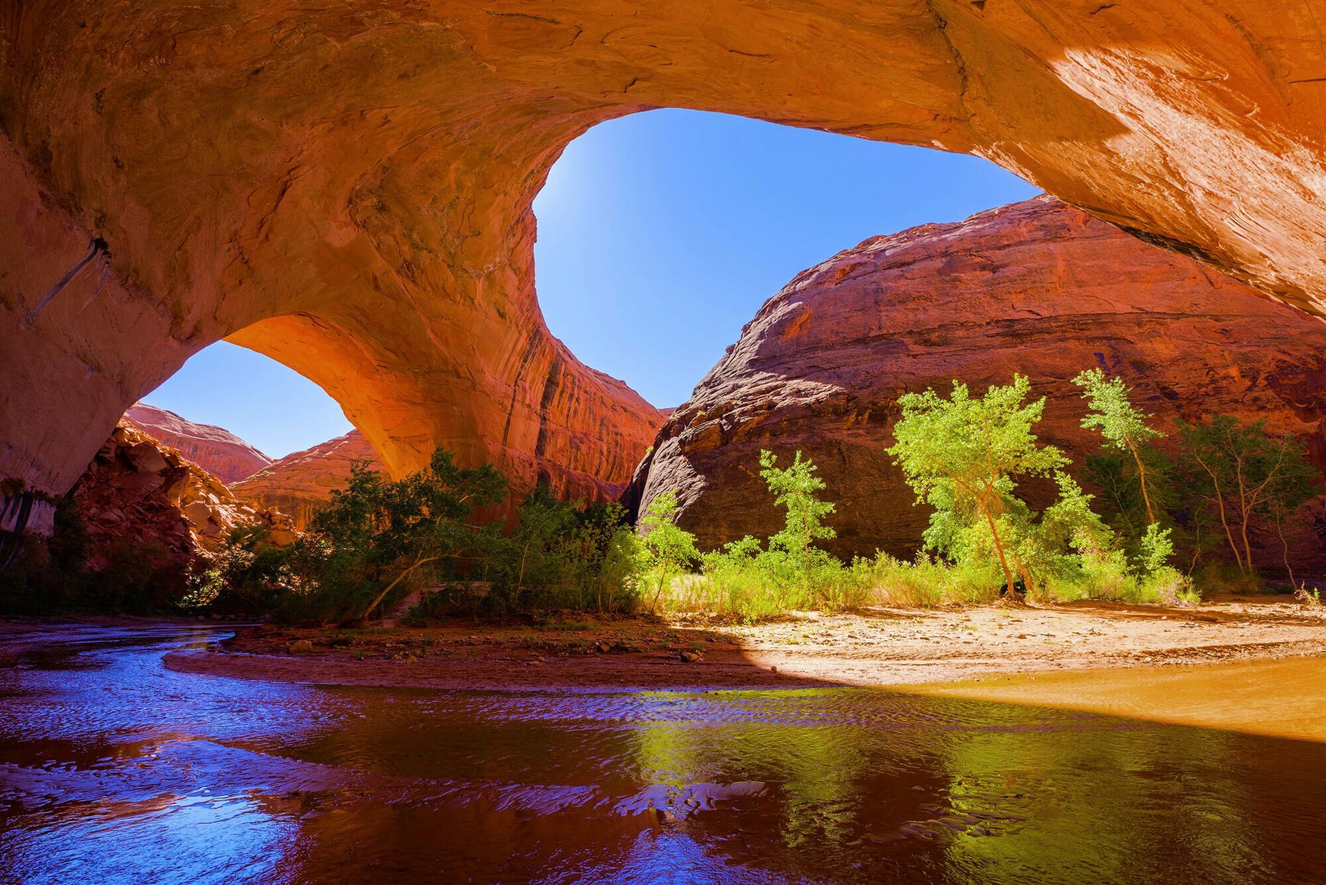 A natural sandstone arch frames a lush green riverbank and canyon walls under a clear blue sky.