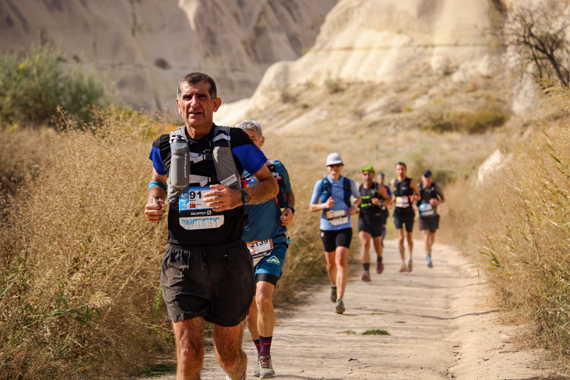 Trail runners race along a dusty desert path beside rocky hills.