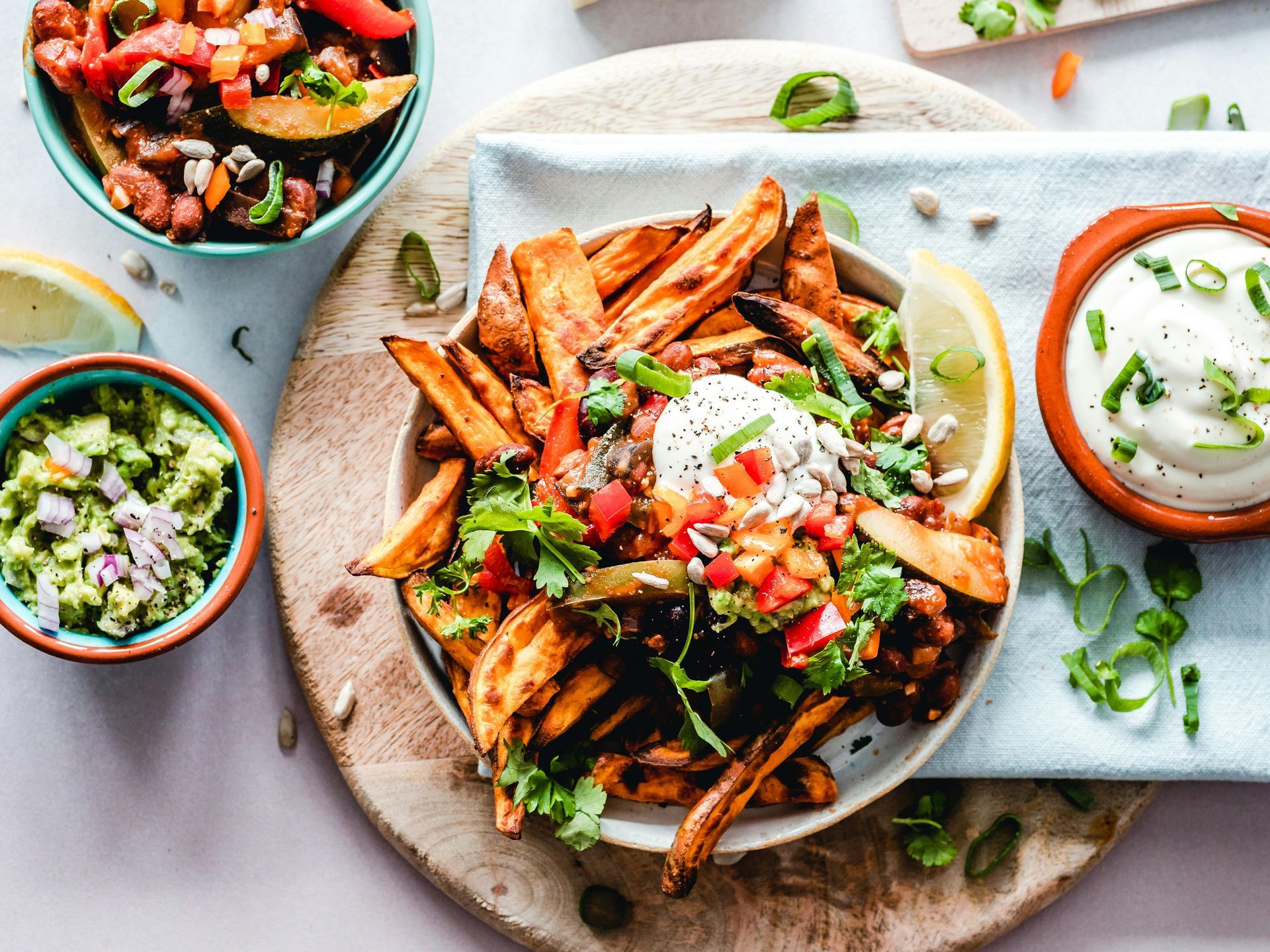A bowl of loaded sweet potato fries topped with beans, peppers, and crema, served with sides of guacamole and cream sauce.