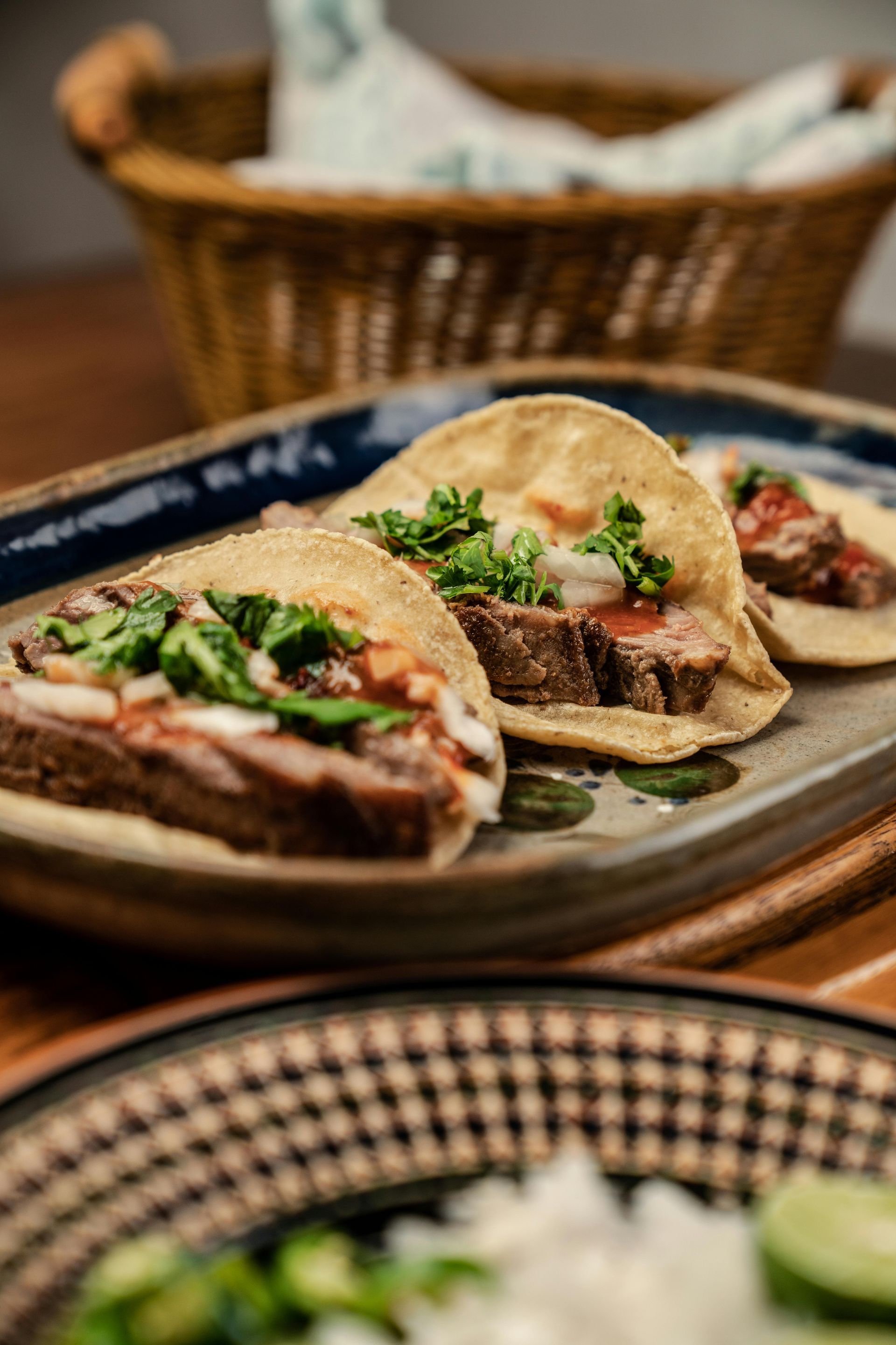 Three street tacos topped with meat, onion, and cilantro on a rectangular platter, with a basket of tortillas nearby.