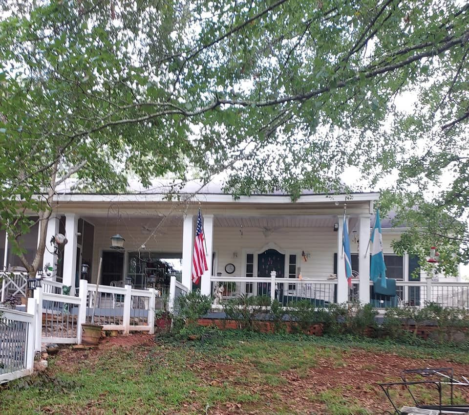 White house with porch, American and Guatemalan flags, surrounded by trees and a white picket fence.