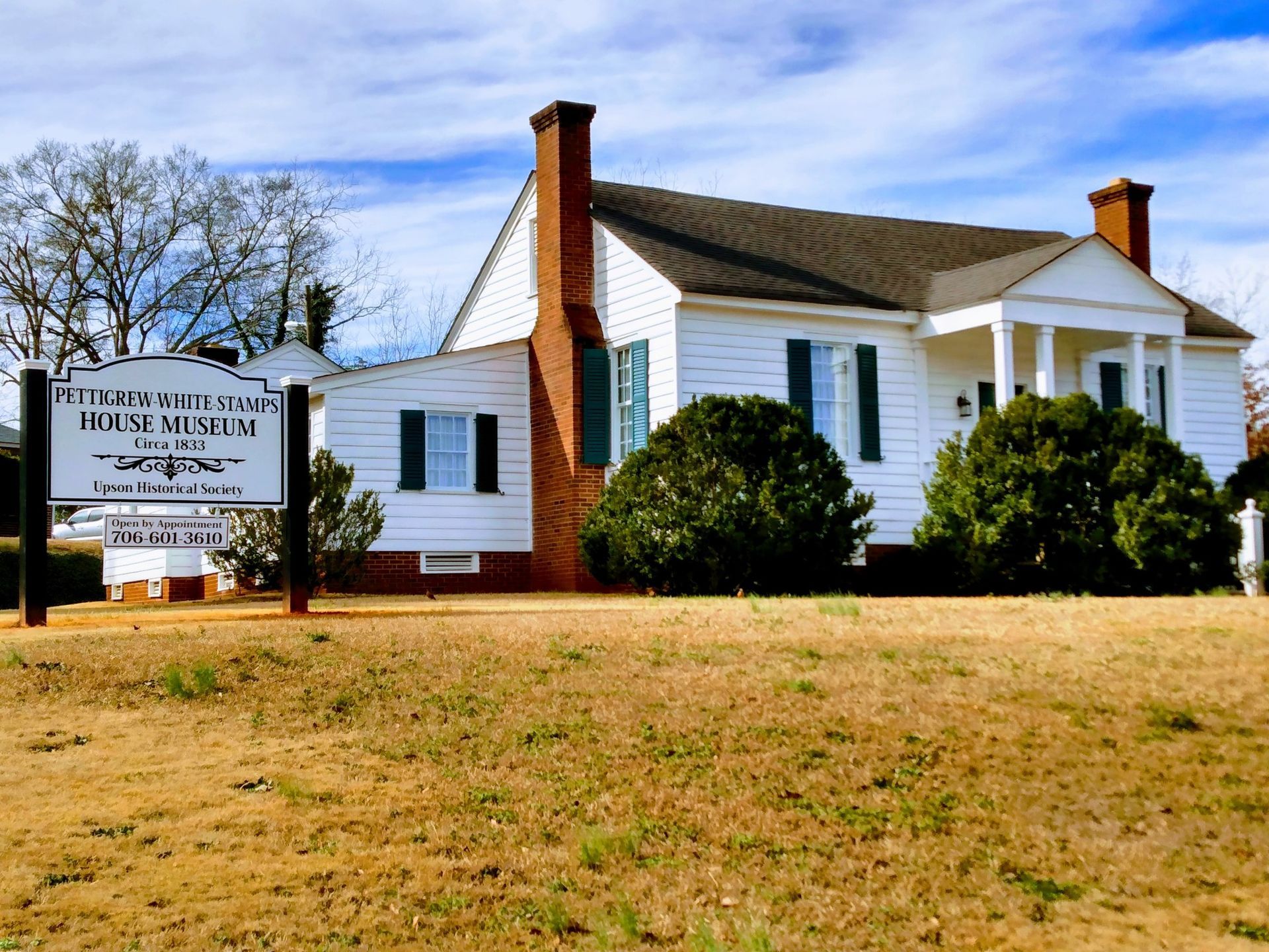 White house with a sign for the Pettigrew State Park's House Museum, green shutters, and a brown yard.