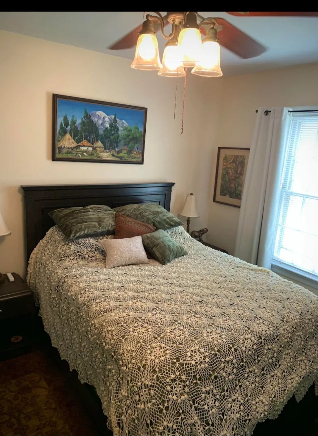 Bedroom with a dark headboard, bed covered in a cream-colored woven blanket, art on the walls, and a window.