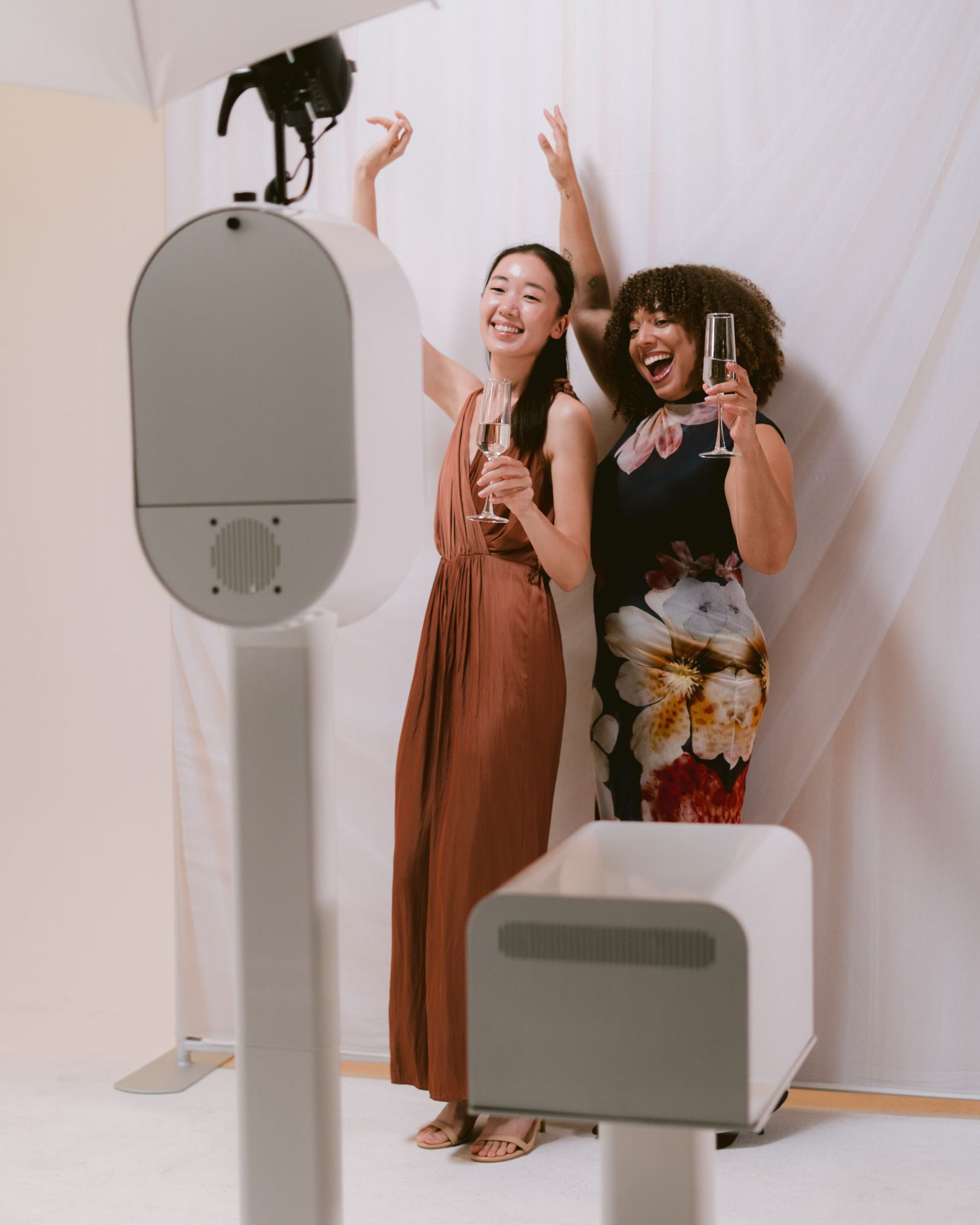 Two guests in formal dresses laughing with champagne in front of the Be You Luxe Booth