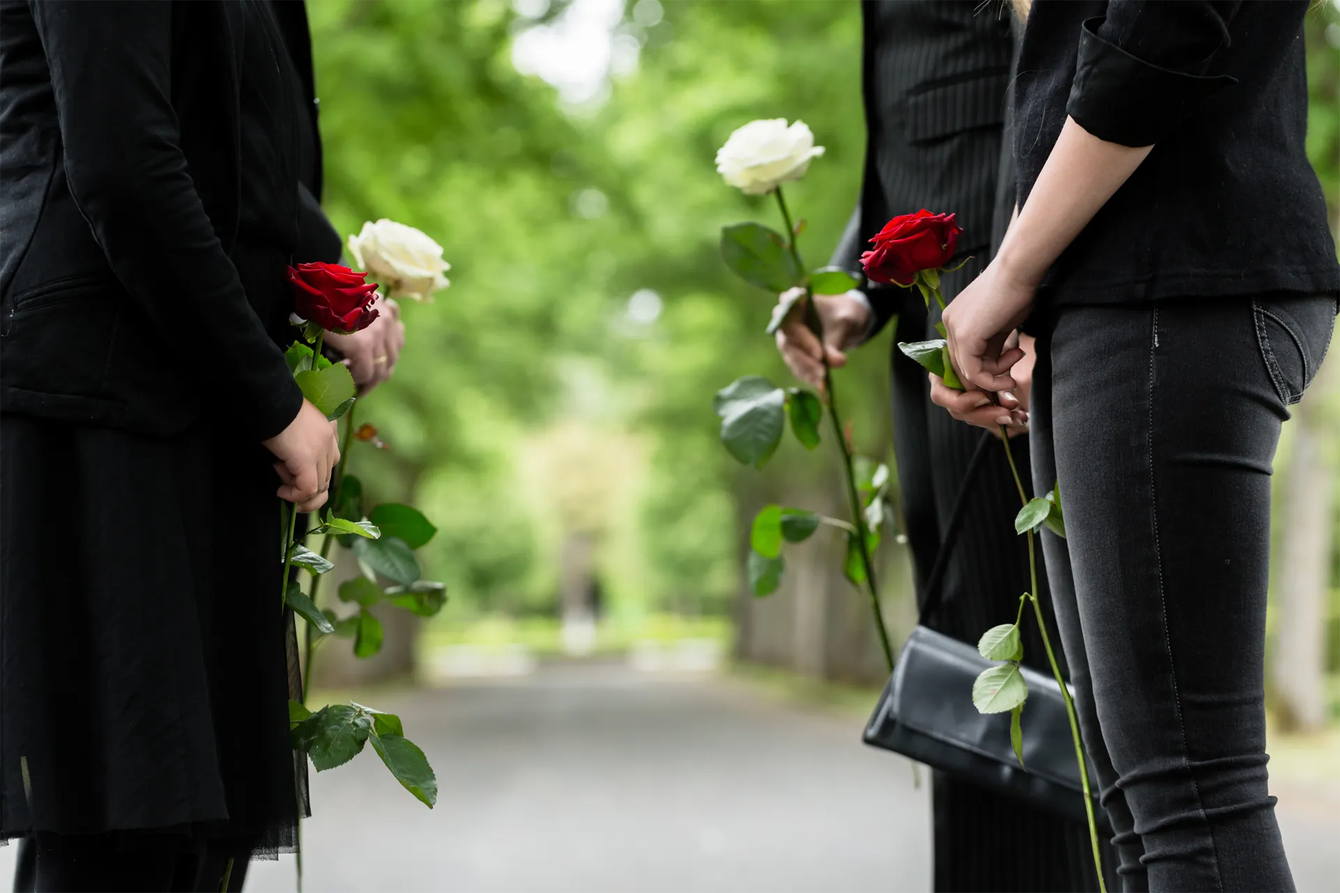 Two people holding roses stand at a memorial path with a blurred green background.