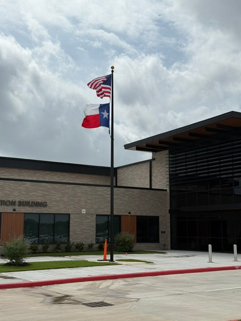Entrance of a public building with Texas and U.S. flags, cloudy sky, and covered walkway