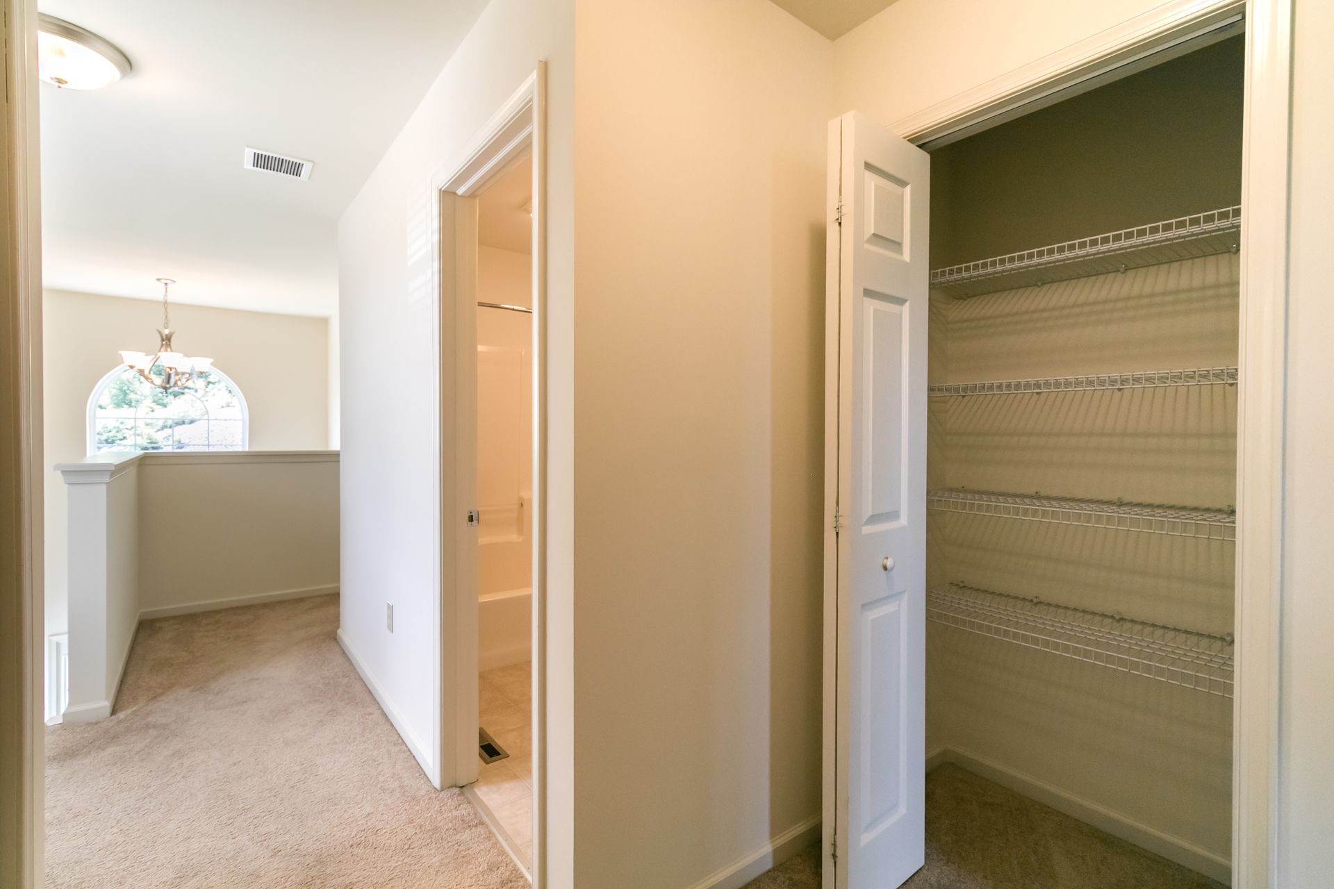 Hallway with open closet, door, and railing leading to a light-filled room; neutral tones.