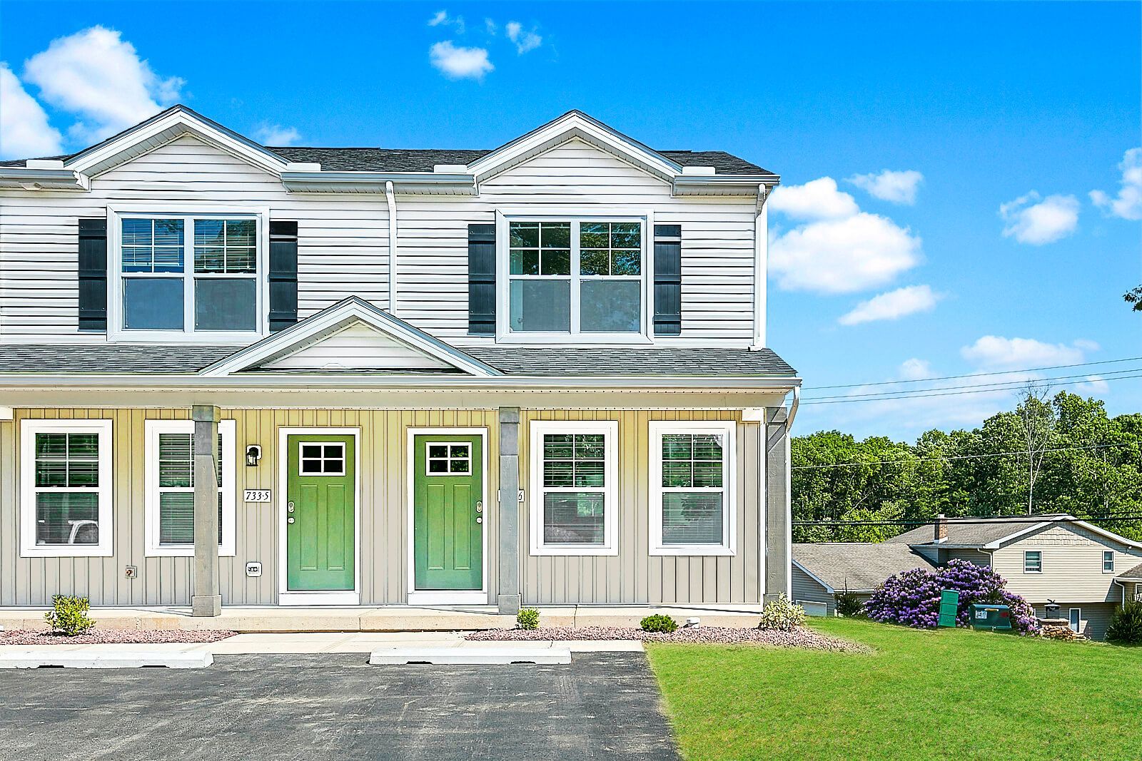 Two-story townhouses with light siding and green doors against a blue sky, green lawn in front.