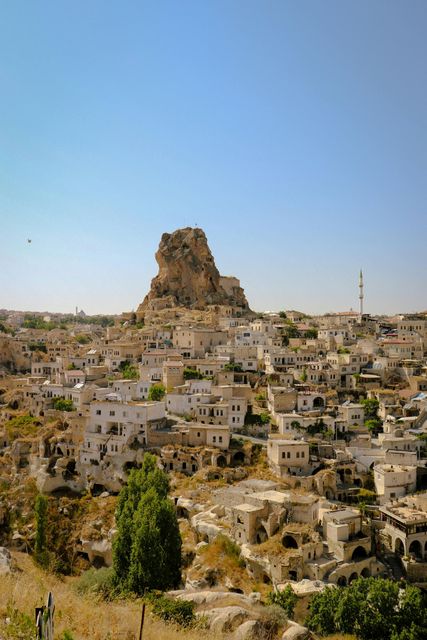 Cittadina collinare con edifici in pietra beige ai piedi di uno sperone roccioso sotto un cielo azzurro limpido.